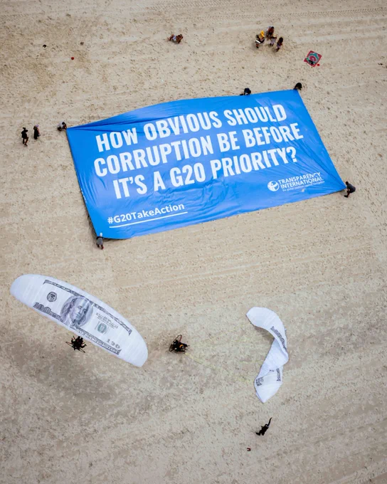 Paragliders with canopies customised with US$100-bill designs flying in over a banner which reads: “How obvious should corruption be before it’s a G20 priority?”