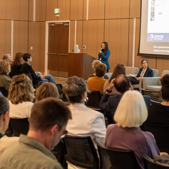 Image Description: A speaker with a microphone stands at a podium in front of an audience. Louisa Bukiet, a white woman with brown hair pulled back, sits to the right of the podium in front of a projector. They are in a conference room with wood-paneled walls.
