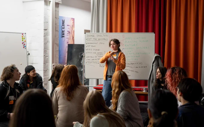 A woman pitching a game idea with a whiteboard behind her in front of a group of people.
