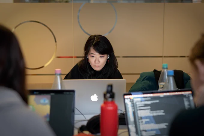 A woman sitting at a table, looking at a Macbook screen in concentration while being surrounded by other hardware and waterbottles.