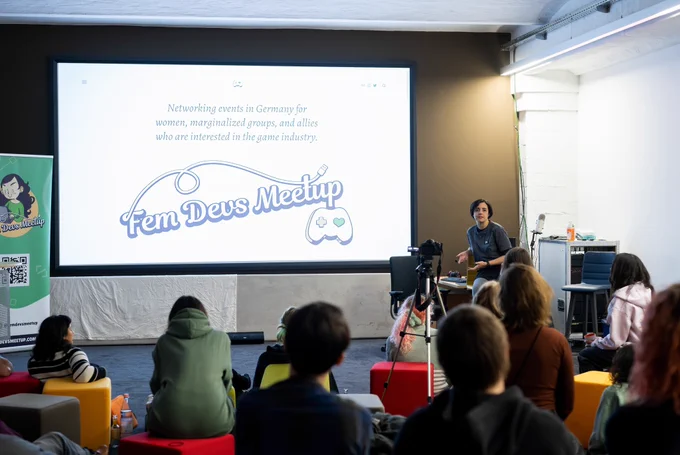 A woman holding a presentation about the FemDevsMeetup initiative on a projector screen in front of a group of people.