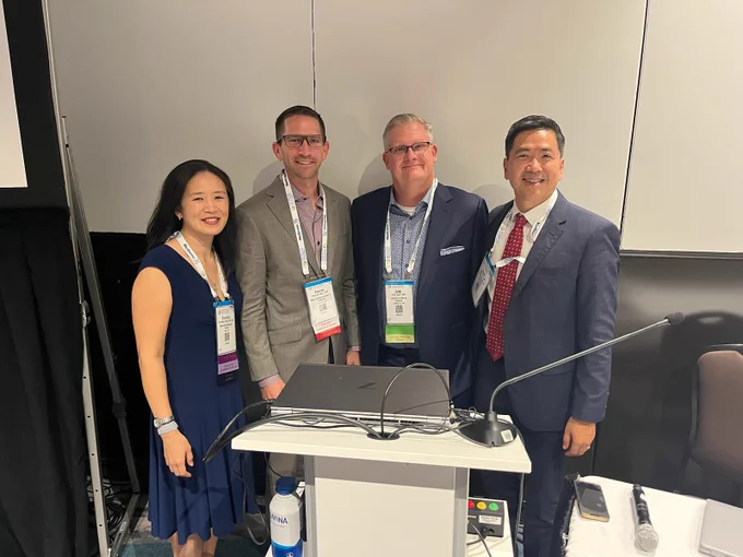 Four professionals, smiling, standing side by side behind a podium at a conference, wearing conference badges and business attire.