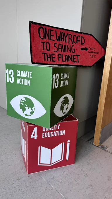 A red road sign with the words “One way road to saving the planet ➡️ Paris Agreement 1.5° C” and two cubes showing SDGs 13 and 4
