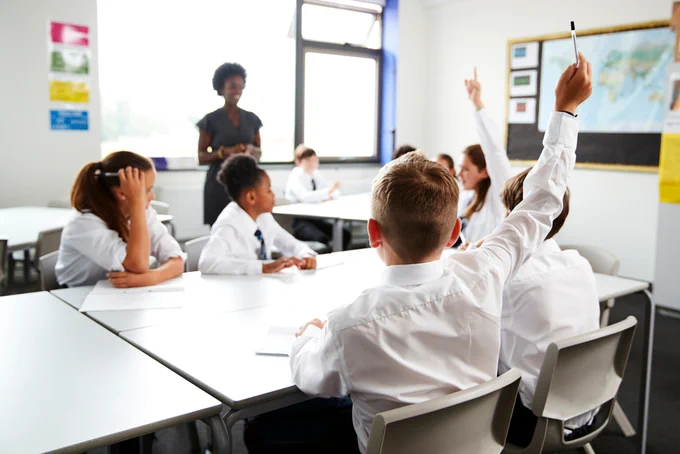 Image of students in the classroom with two students with their hands up and the teacher in the background.