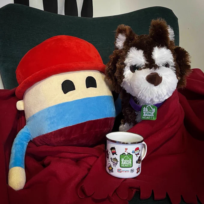 Plushies of Tyke and Bonnie the dog from Pine Hearts sit on an arm chair. They're wrapped in a red blanket and have a Pine Hearts mug sitting between them.