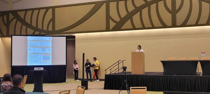 Me presenting my poster, Examining Mainstream News Media Narratives on Youth Online Safety, on a stage in a conference hall