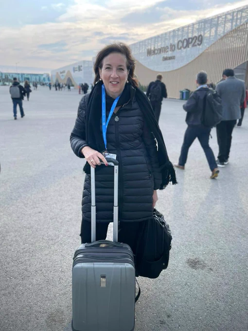 PRM Acting Assistant Secretary Marta Youth stands with a suitcase in front of a building with signage reading, "Welcome to COP29."