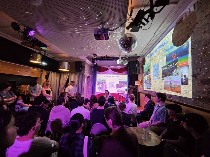 A photograph from game-a-week party. A crowded room watching a presentation. There's mood lighting and a disco ball.