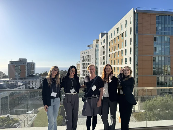 Five female presenting individuals standing together on a balcony with buildings and a clear sky in the background. They are all smiling and wearing name tags.