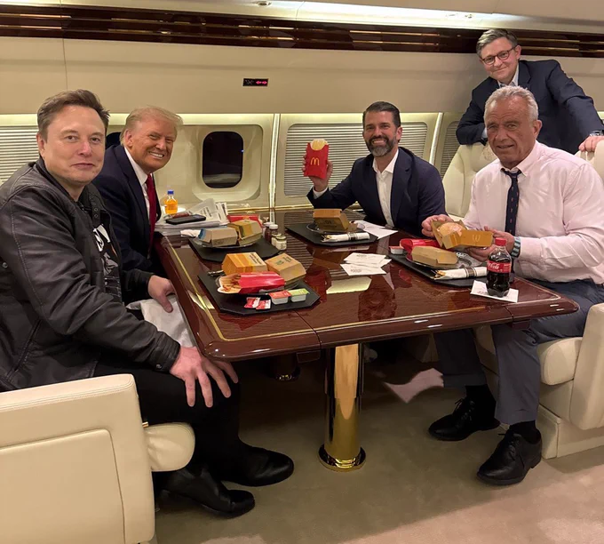 Trump, one of his doofus sons, Kennedy and Trump's emotional support billionaire posing at a table with a cold McDonalds