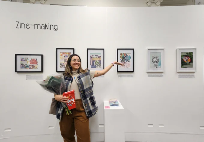 A woman wearing brown trousers, a beige blouse and checked gillet is standing in front of a white gallery wall and holding flowers and chocolate. The person is pointing to frames on the wall and smiling. A wall sticker reads 'zine-making'. There are six frames showcasing artwork on the wall.