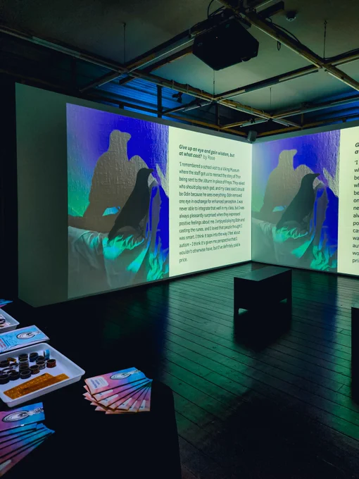 A room whit a slideshow projected on to a wall. The projection shows the picture of a crow in blue and green light. Next to the picture, there is some text and the caption 'Give up an eye and gain wisdom, but at what cost? By Rose'
There is a bench facing the wall with the projection. To the left on the foreground, there is a table containing colorful booklets and craft materials.