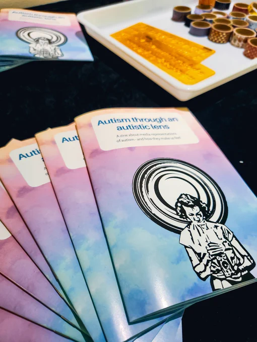 Colorful booklets displayed on a table. On the cover, there is a black and white illustration of a woman holding a vintage camera and looking down at it. The title of the booklet is 'Autism through an Autistic Lens - a zine about media representations of autism and how they make us feel'. Behind the booklets, some craft materials are also displayed on the table.