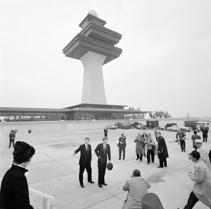 JFK, hat in hand, stands outside the Dulles Airport with Najeeb Halaby. Members of the press take photos.