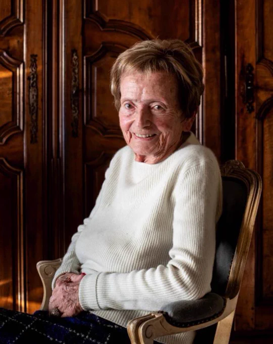 An elderly woman smiling while seated in an ornate wooden chair in a warmly lit room.
