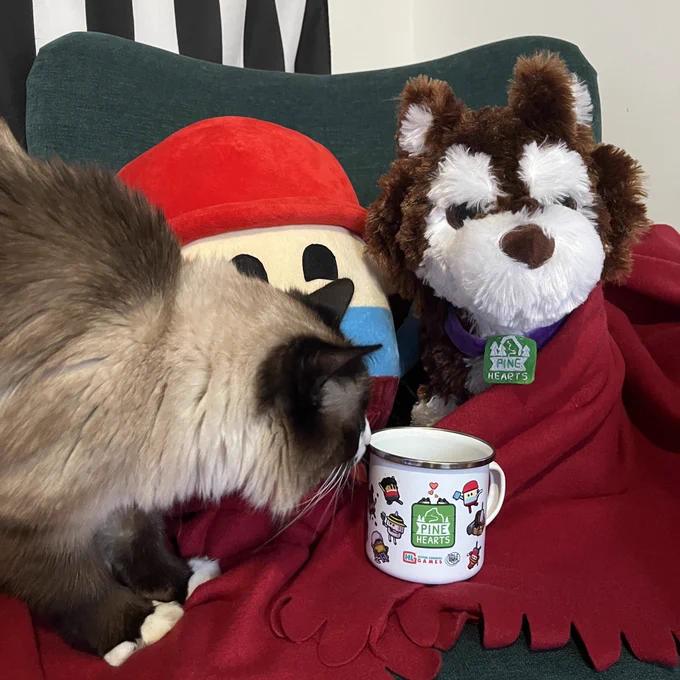 Tyke and Bonnie plushies from Pine Hearts set up on an arm chair wrapped in a red blanket. A seal point ragdoll cat has walked into frame from the left hand side and is sniffing a mug in the centre of the image.