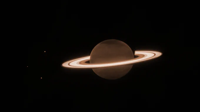 Image has a mostly dark background and at the center is a dark orange brownish circle, surrounded by several blazing bright, thick, horizontal whiteish rings. This is Saturn and its rings. There are three tiny orange dots in the image — one to the upper left of the planet, one to the direct left of the planet, and the lower left of the planet. They are the moons Dione, Enceladus, and Tethys.