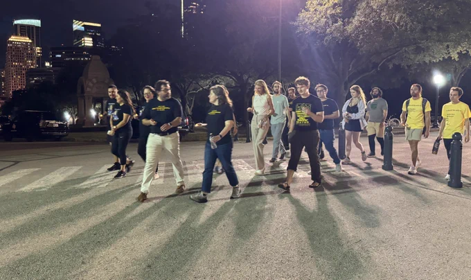A group of people walking around the Texas State Capitol loop, with Downtown Austin buildings in the background.