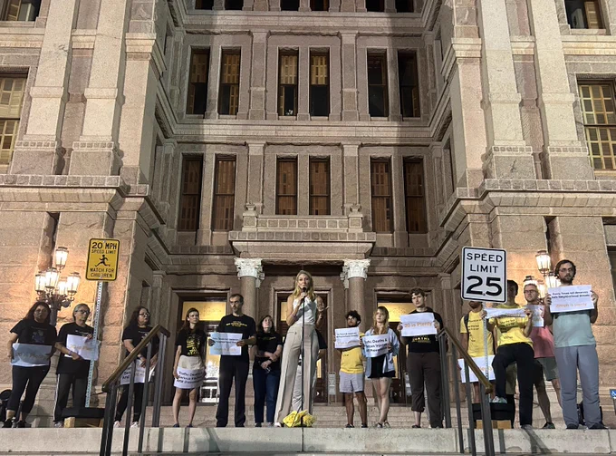 A group of people standing on the front steps of the Texas State Capitol with Jamie White in the center on the microphone and low speed limit signs.