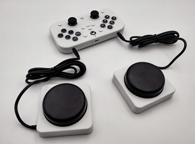 Fairly thick flat white gamepad with black Xbox style buttons and thumb sticks. Rear view has two 3.5mm black cables plugged in either side of the back edge, leading to two medium sized round black raised push buttons in a white black square base with rounded off corners.