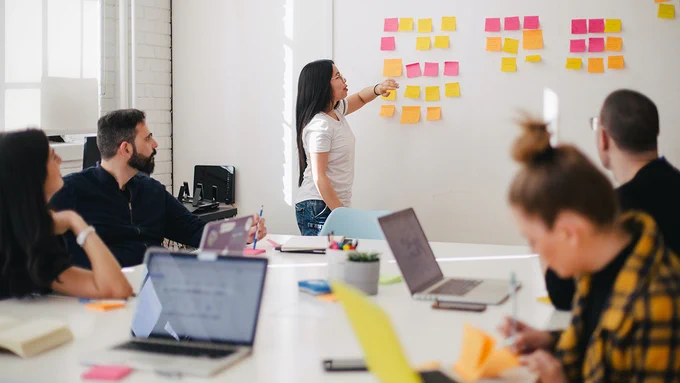 Students in a workshop sat around a table, with one person stood up presenting at a white board.