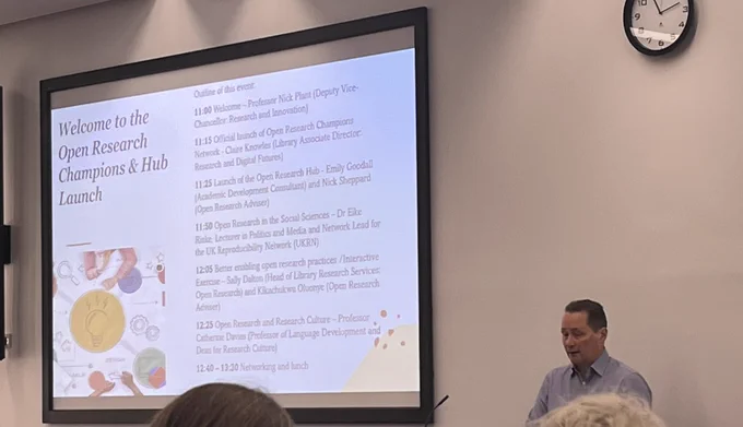 Large digital screen showing a slide about an Open Research event, with the presenter stood to the side of it.