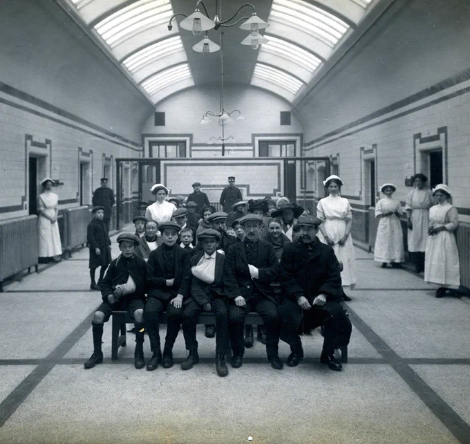 Black and white photo of a group of patients sitting in a hospital waiting room. There are several nurses standing along the side of the waiting room. 