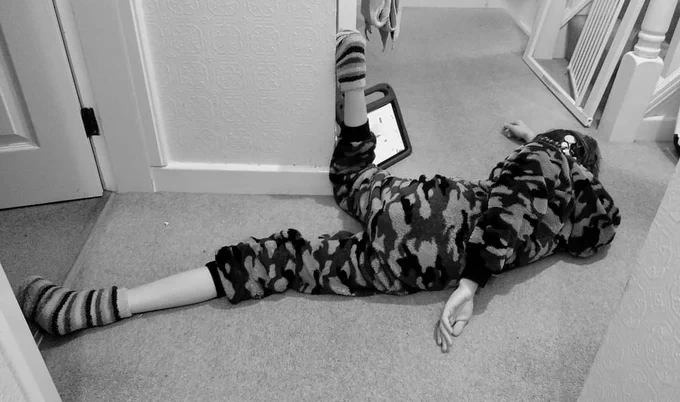 Boy in camp fleece onesie and fleece socks lies on the carpeted floor of a home. He's facing away from the camera looking at an ipad propped up. One leg is at 90degrees from the knee against the edge of a corner of the wall, both arms and lying along the floor - a posture which offers lots of sensory input. 