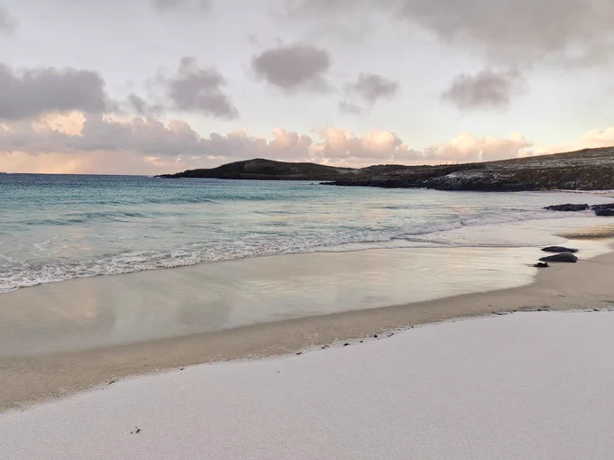 Image shows snow on beach with turquoise sea and orange clouds in background 