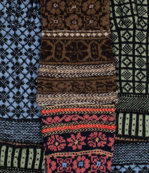 A close up of different knitted gloves and mittens overlaid on one another. Each is knitted with an intricate pattern, one is brown, one is green, one is blue and one is red.  