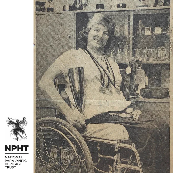 A black and white newspaper image of Gwen Buck, aged 43, seated in her wheelchair in front of a wooden display cabinet at home, surrounded by medals and trophies. She is smiling.