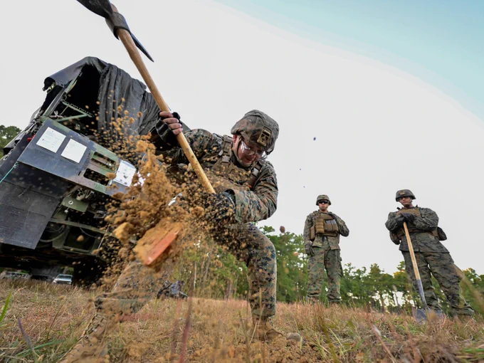 Marine Corps Reservist digs into the ground with a shovel during a field training exercise, as two fellow Marines observe nearby. A tactical vehicle is visible in the background, emphasizing the operational setting. (DoD photo by Nicholas Pilch)