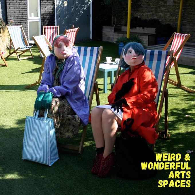 Two people wearing masks sitting on stripey deck chairs in the garden. They have on brightly coloured rain jackets, wigs, gloves and scarves, and one holds a blue and white stripey bag in front of their legs.