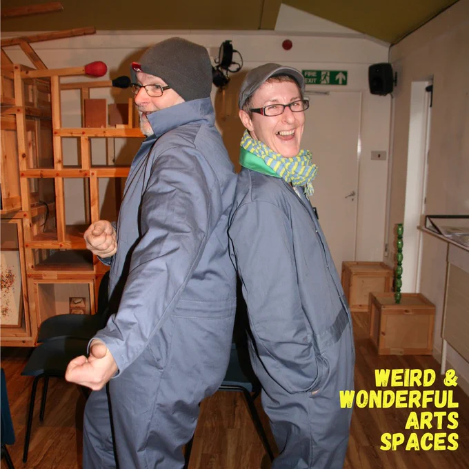 Bob and Sarah smiling and standing back-to-back in grey overalls and hats in front of the lending library space.