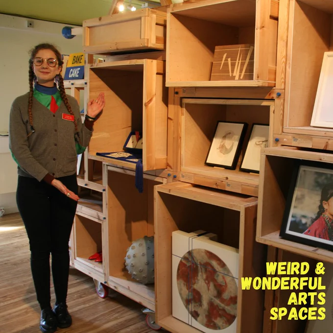 Pennelope, an art library volunteer, stands in front of the display with arms outstretched, in the grey, green, blue and red uniform.