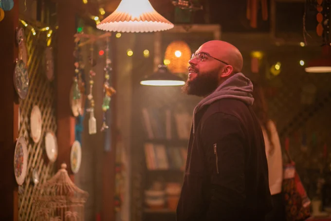 A man wearing a grey hoodie and black jacket looks up at a warmly light courtyard space with hanging decorative plates.