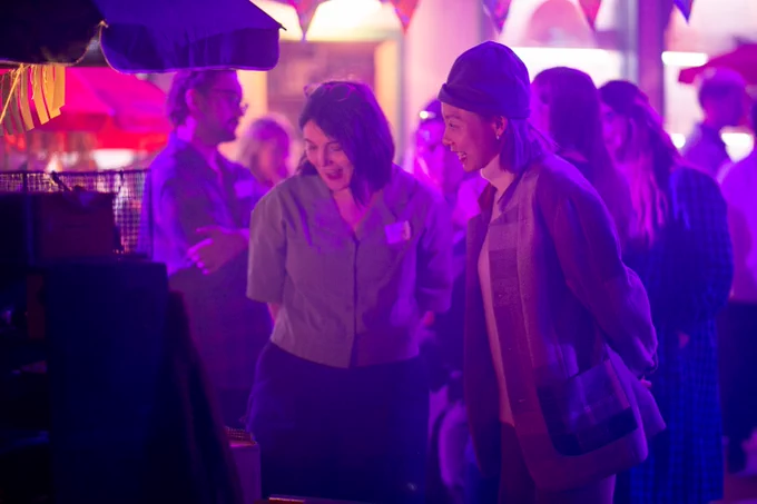 Two people look excitedly at an instillation. They are lit by a purple light and there is bunting above them. 