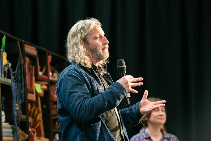 Peter Higgin speaks on stage. He is wearing a blue jacket and holding a microphone. There is a bookcase behind him.