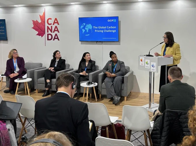 A panel discussion at an event focused on "The Global Carbon Pricing Challenge," as indicated on the screen behind the speakers. Five women are involved: one standing at a podium speaking and four others seated in chairs on the stage. Attendees are seated in the foreground, listening, some with headphones for translation. 