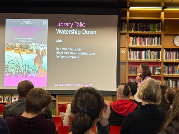 Photo from the back of an audience in a library, with a view of a white woman with brown hair (me) gesticulating towards a screen that says: Library Talk
Watership Down with
Dr. Catherine Lester
Nigel and Klive Humberstone 
Dr Sam Summers 