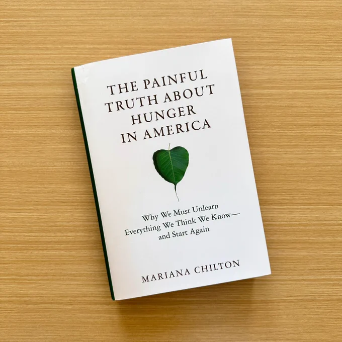 A book titled "The Truth About Hunger in America" by Mariana Chilton, displayed on a wooden surface. The cover features a minimalistic design with a green leaf graphic.