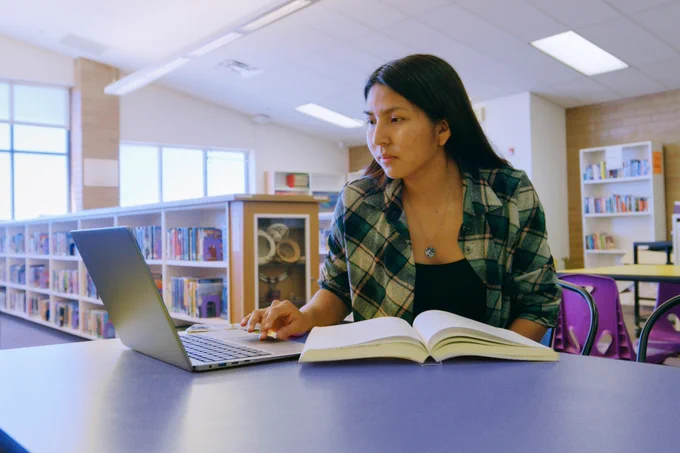 A young Indigenous woman sits in a library, focused on her laptop, with an open book in front of her. Bookshelves and bright natural light fill the background.