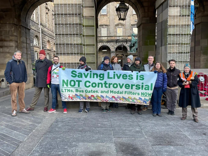 Group of people in warm clothes standing in front of Edinburgh City Chambers with a banner reading:
Saving lives is NOT controversial! LTNs, bus gates and modal filters NOW!