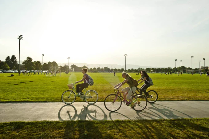 Three persons riding a bike on a trail on the C S U campus. Intramural fields are visible in the background.