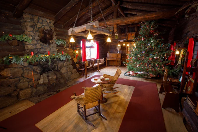Interior of a a log-cabin style room decorated for Christmas with a large tree adorned with lights and ornaments and garland on the chimney hearth. Two wicker sleigh chairs face the tree, and sunlight streams through the window.