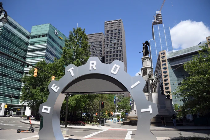 A view of a cityscape featuring an arch with the word "DETROIT" at the forefront, a statue, and several modern buildings under a clear sky.
