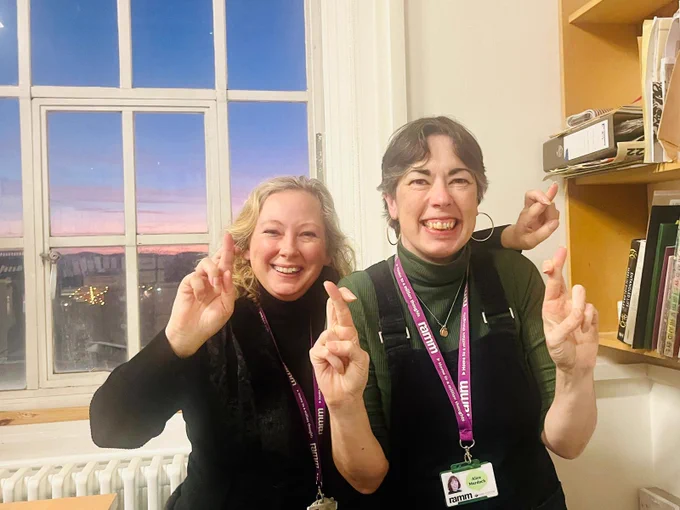 two women standing in an office in front of a window showing a sunset sky, holding up crossed fingers
