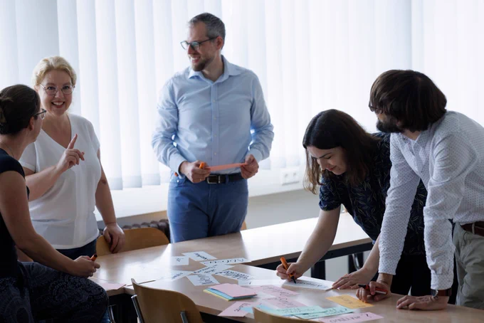 Fünf Personen arbeiten in einem Seminarraum an einem Tisch zusammen. Einige der Personen stehen, andere beugen sich über den Tisch, auf dem bunte Notizzettel und Papierstreifen mit Text verteilt sind. Im Hintergrund sind helle vertikale Jalousien zu sehe