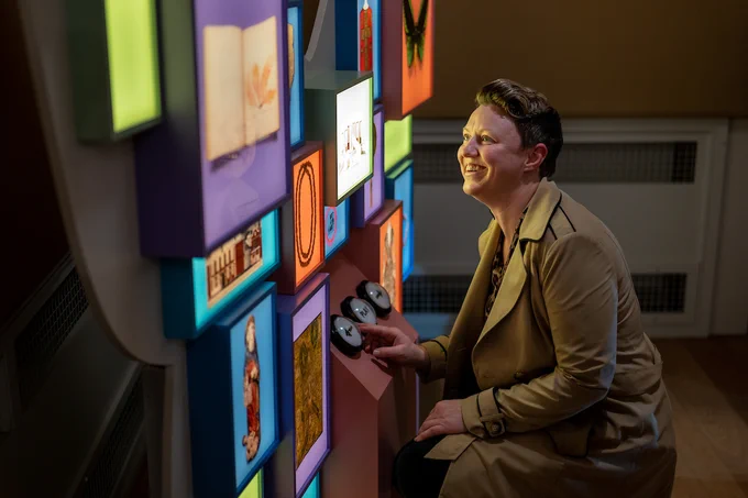 a person in a beige jacket with short hair smiling and kneeling down in front of a butterfly shaped museum interactive
