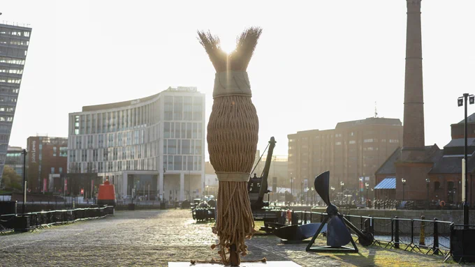 LR Vandy's roped sculpture Dancing in Time: The Ties That Bind Us stands on Albert Dock. Sunlight bursts from behind it.